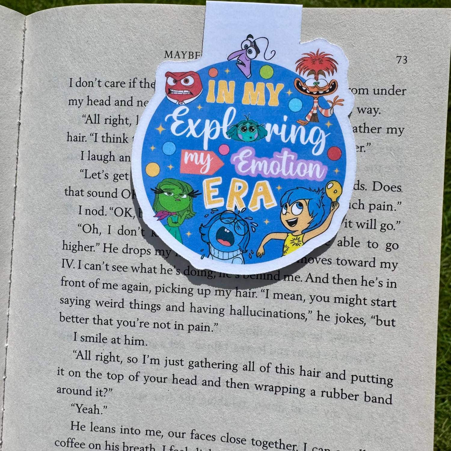A round blue book bookmark with colorful cartoon characters and the text 'IN MY Exploring my Emotion ERA' placed on an open book outdoors on grass.